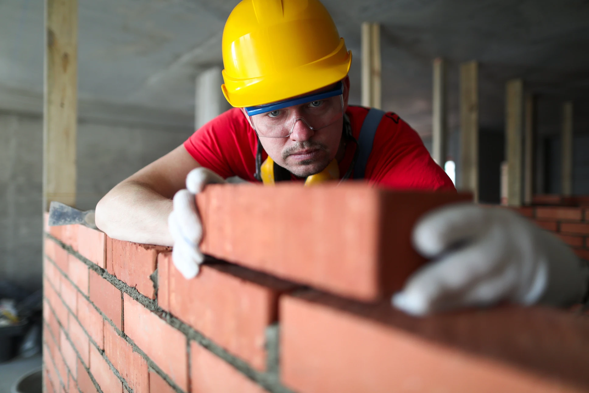 Ouvrier en bâtiment posant des briques rouges sur un mur en construction, portant casque de chantier jaune, lunettes de protection et t-shirt rouge